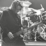 A black and white, mid-shot photo of Chris Gaylord, the bassist for Wage War, playing a black bass guitar on stage. He has long, dark hair covering his face as he looks down at his instrument, deeply engrossed in his performance. He is wearing a dark, possibly collared, long-sleeved shirt. In the blurred background, the drummer, Stephen Kluesener, is visible behind his drum kit, which features a bass drum with a white, geometric star-like logo. The image is lit with bright stage lights, creating strong contrasts.