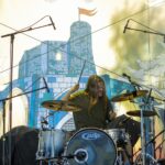 Drummer of Ringscabs performing live in front of a large hanging displaying the artwork of the festival with a blue drawing of Gisors' castle.
