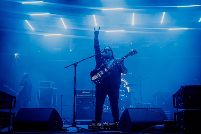 Man holding a guitar on stage throwing up the horns with his band