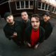 A high-angle, wide-angle fish-eye lens photograph of the four members of the band Vicious Rain. They are standing outdoors on pavement in front of a brick building with several windows. The band members wear dark jackets and hats, with the central figure wearing a vibrant red t-shirt. They look directly up into the camera with serious expressions. Several members have visible tattoos and facial piercings.