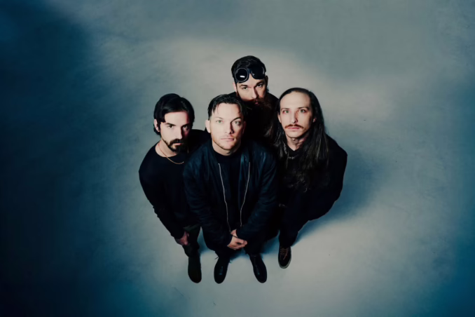A professional promotional group portrait of the metalcore band LEVELS. The four members are posed in an urban outdoor setting against a neutral stone wall. They are dressed in contemporary streetwear—hoodies, flannels, and black denim. From left to right: the bassist, guitarist, vocalist Kolby Carignan in the center, and the drummer. The lighting is soft and natural, emphasizing a serious and cohesive band identity.