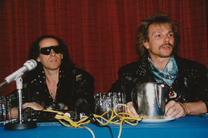 A candid 1989 photograph of Scorpions members Klaus Meine and Rudolf Schenker seated at a press conference table with a blue tablecloth. To the left, vocalist Klaus Meine wears dark oversized sunglasses and a studded black leather jacket. To the right, guitarist Rudolf Schenker sports his signature mustache and a leather jacket with a patterned blue scarf. Microphones and a silver pitcher sit on the table in front of them against a backdrop of heavy red curtains.