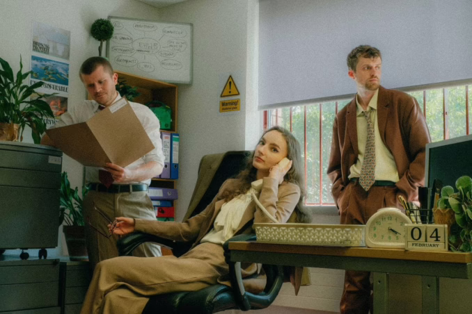 Two males standing, one female sitting in a chair by a desk