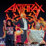Joey Belladonna singer of Anthrax performing live in front of a display of their album cover, Cursum Perficio. He's making a crushing hand gesture.