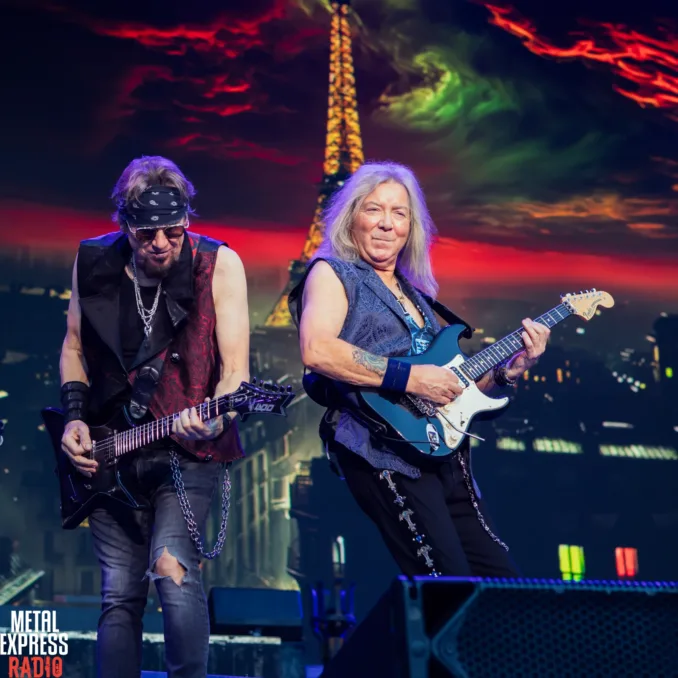 Adrian Smith and Dave Murray, guitarists of Iron Maiden playing side by side during live performance. They are wearing subtly matching waistcoats. The background features a view of Paris and the Eiffel tower caught in a menacing storm...although the performance was in Lyon and the French fans were grumpy about it.