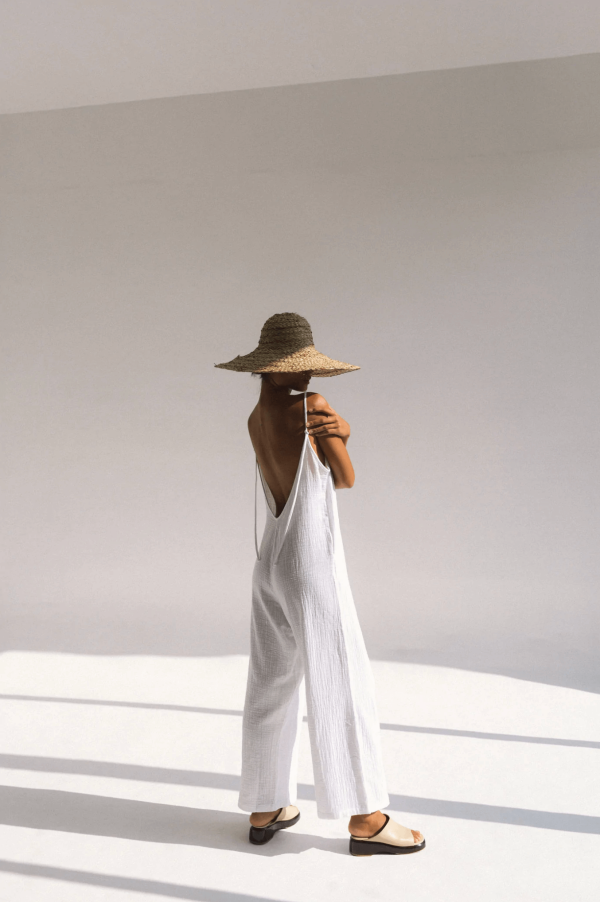 A woman wearing the Flo Jumpsuit in White by Bahhgoose, styled with a hat, showing the open back and tie straps