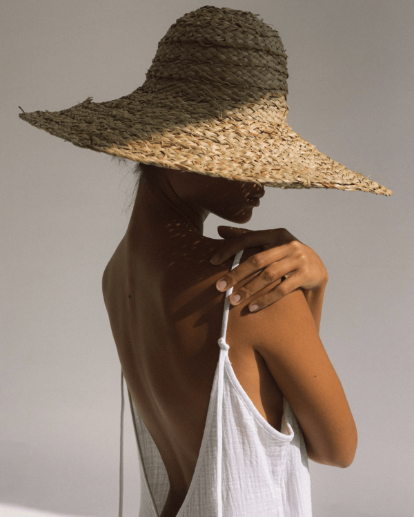 A close up of a woman wearing the Flo Jumpsuit in White by Bahhgoose, styled with a hat, showing the open back and tie straps