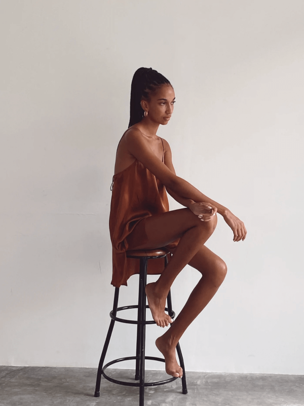 a woman sitting on a chair while wearing the Jai Dress in Coco by the brand Bahhgoose, showing the relaxed fit and soft silky fabric of the dress