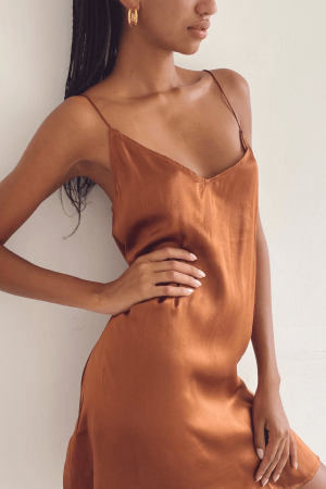 a woman leaning against a wall while wearing the Jai Dress in Coco by the brand Bahhgoose, showing the relaxed fit and soft silky fabric of the dress