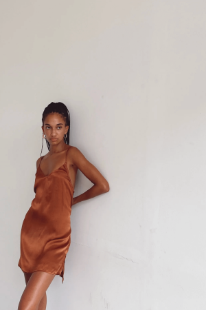 a woman leaning against a wall while wearing the Jai Dress in Coco by the brand Bahhgoose, showing the relaxed fit and soft silky fabric of the dress