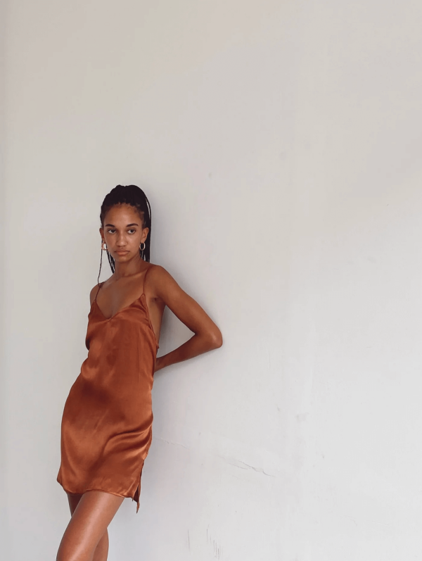 a woman leaning against a wall while wearing the Jai Dress in Coco by the brand Bahhgoose, showing the relaxed fit and soft silky fabric of the dress