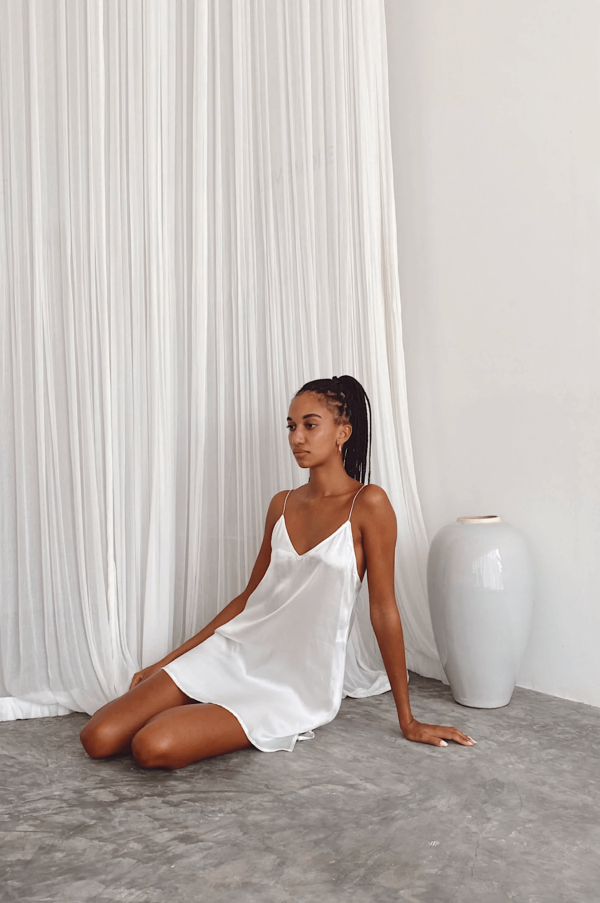 a woman sitting down while wearing the Jai Dress in White by the brand Bahhgoose, showing the relaxed yet refined look of the dress