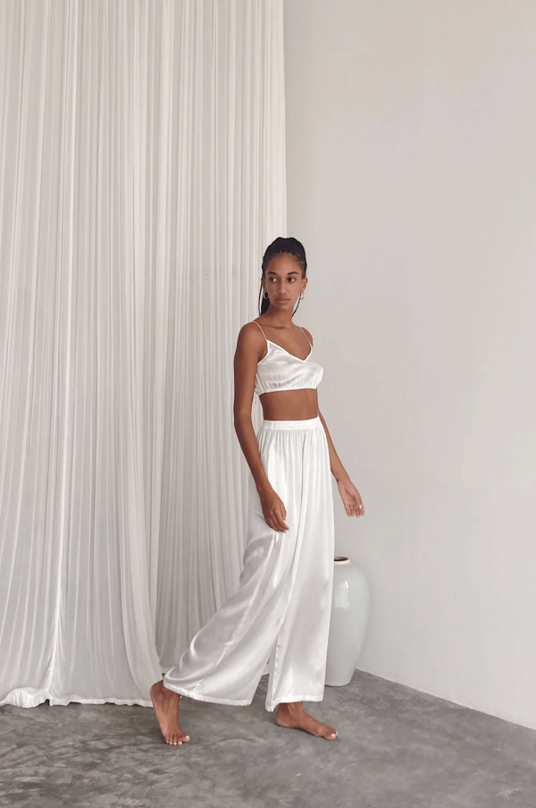 a woman wearing the Jai Pants in White by the brand Bahhgoose, showing the relaxed fit and the silky fabric