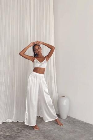 a woman wearing the Jai Pants in White by the brand Bahhgoose, showing the relaxed fit and the silky fabric