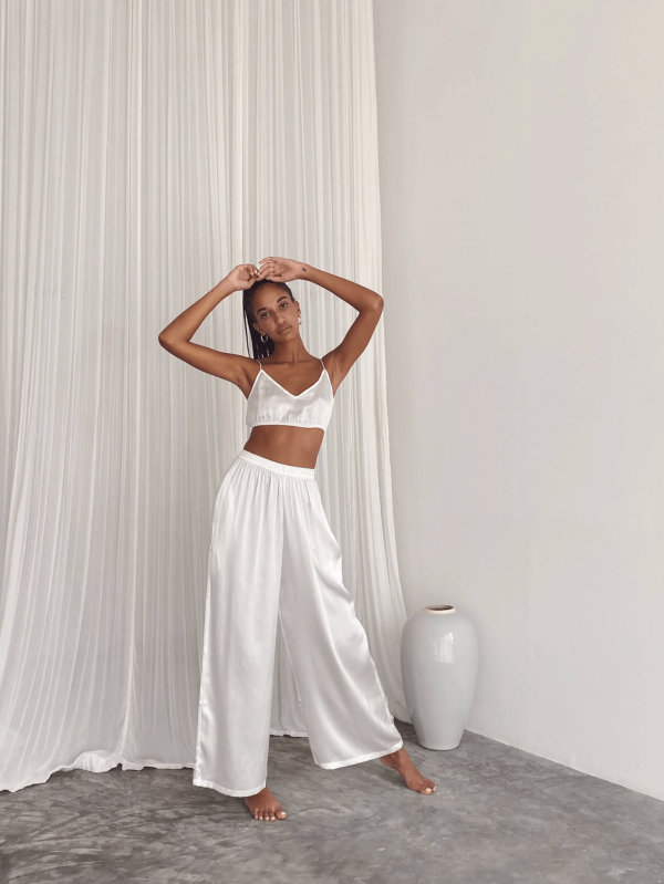 a woman wearing the Jai Pants in White by the brand Bahhgoose, showing the relaxed fit and the silky fabric
