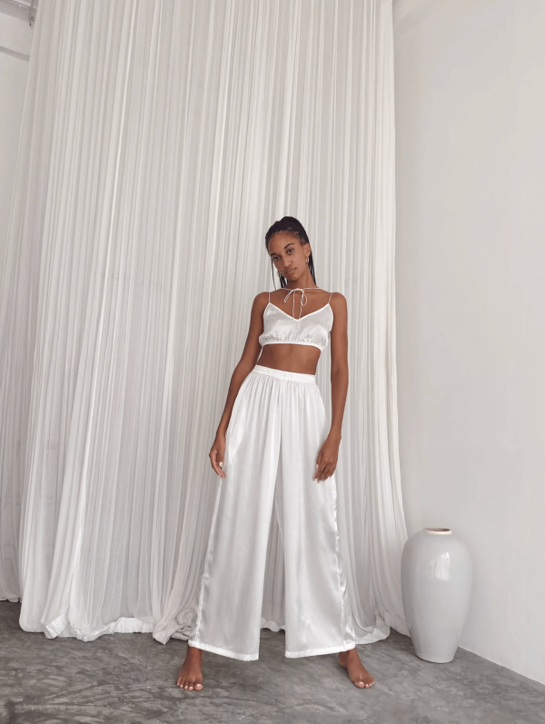 a woman wearing the Jai Pants in White by the brand Bahhgoose, showing the relaxed fit and the silky fabric