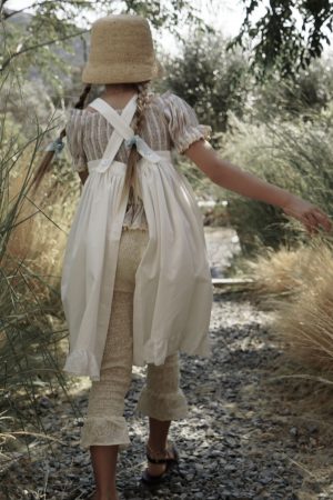 girl wearing the Frida Apron Dress in Cotton D'éte fabric, paired with the Zadie Pants in Creme Broderie & the Sirene Blouse in Sicilian Stripe fabric by the sustainable brand House of Paloma, curated by Morsel Store