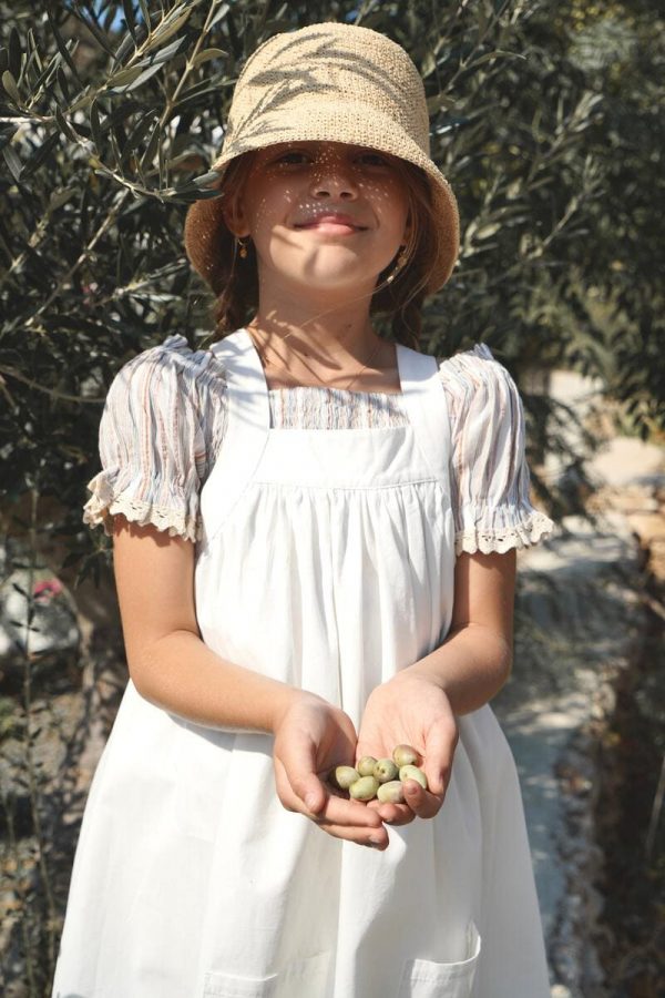 girl wearing the Sirene Blouse in Sicilian Stripe fabric, paired with the Frida Apron Dress in Coton D'été and a raffia hat, by the sustainable brand House of Paloma, curated by Morsel Store