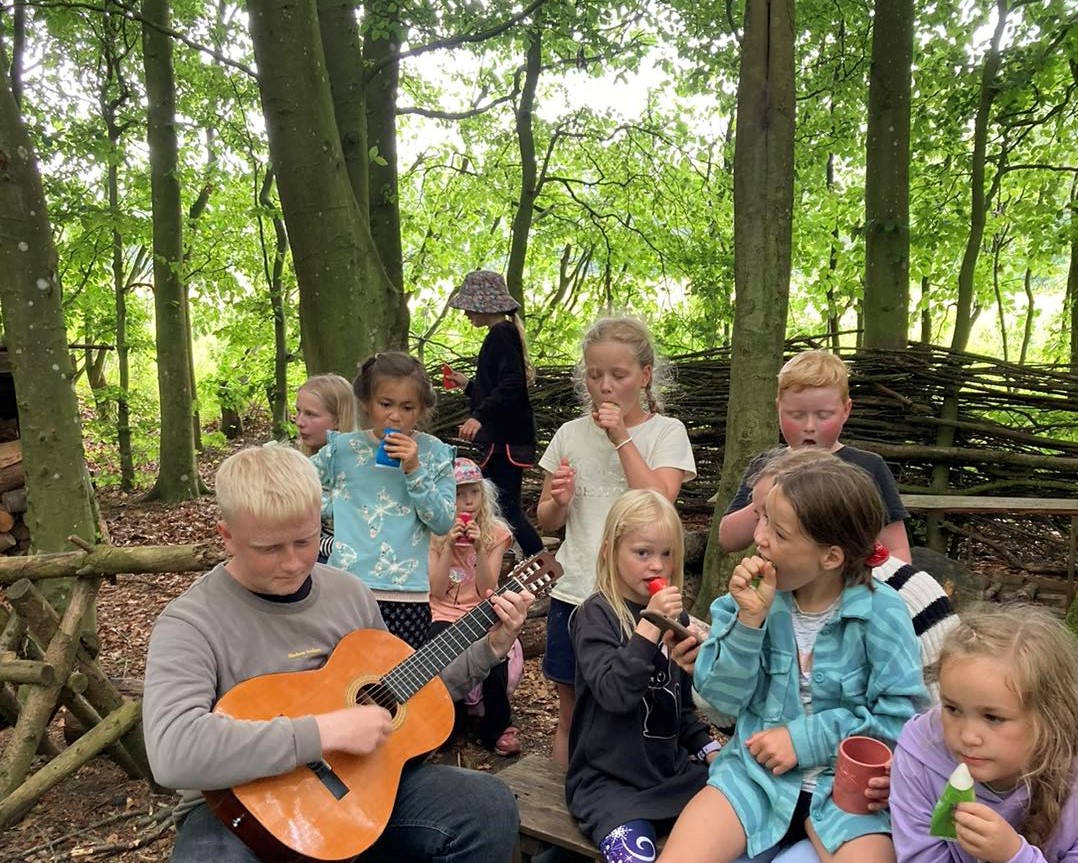 Stor dreng spiller på guitar for sfo børn i den grønne skov ved friskolen