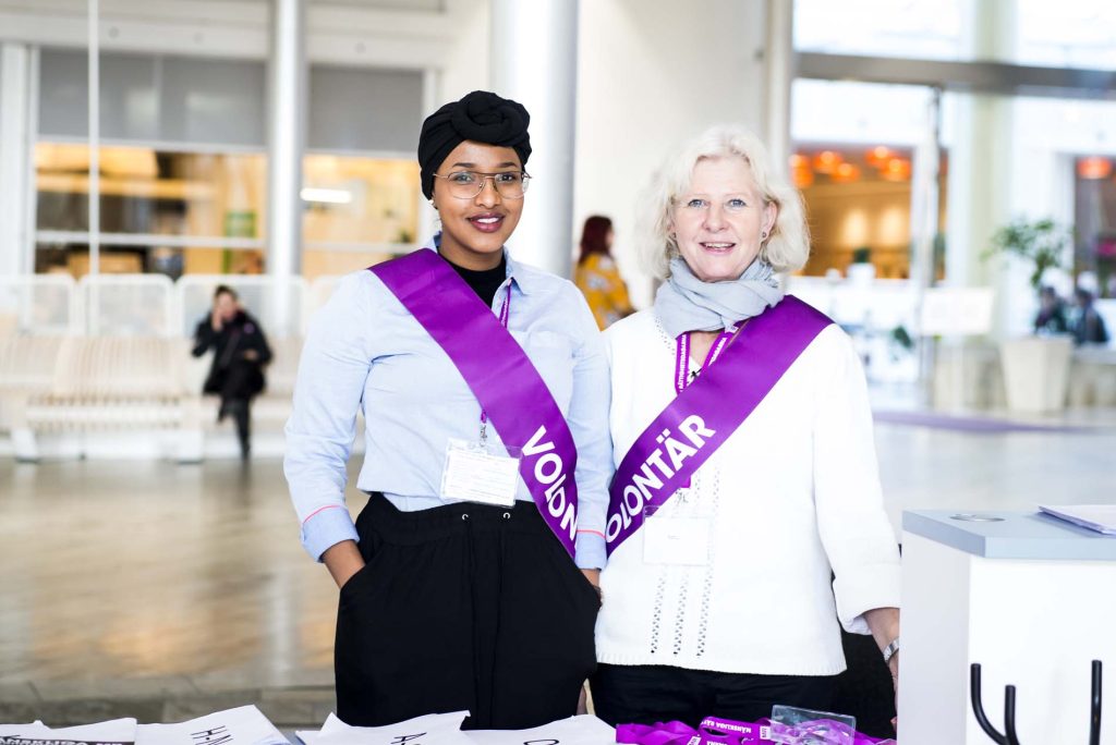 Två volontärer, en ung och en äldre kvinna står brevid varandra. De bär lila band där det står "volontär".