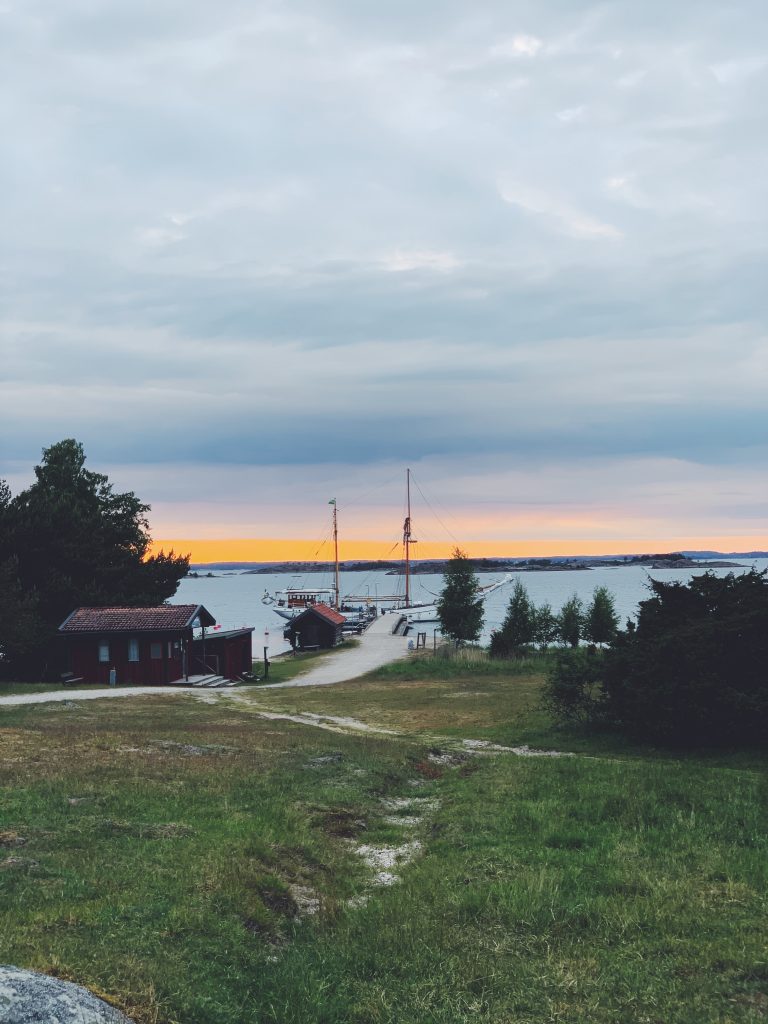 Ångbåtsbryggan från land. Ett vackert segelfartyg har lagt till vid bryggan med en bakgrund av ett apelsinfärgat sken i skyn.