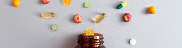 Vitamin pills and capsules alongside apples, bananas, lime and oranges coming out of a medicine jar, nutrition concept