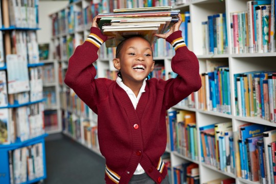 Schoolgirl smiling and balancing stack of books on the head at library