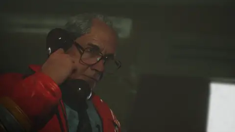 The Chinese Room Screenshot shows a man with large glasses holding an old rotary phone handset to his ear in a dimly lit office. He wears a grave expression as he listens to the person on the other end.