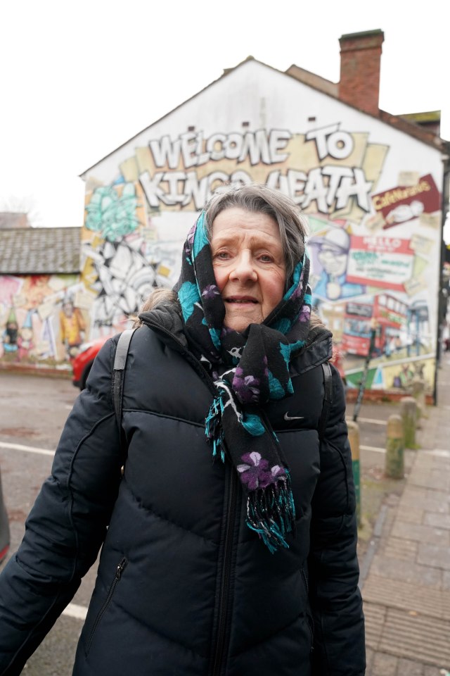 An elderly woman wearing a black puffer jacket and patterned scarf stands in front of a mural that says "Welcome to Kings Heath."