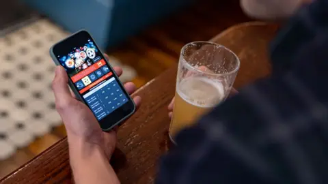 Getty Images Close-up on a man gambling online using a mobile app on his cell phone while drinking beer at the pub