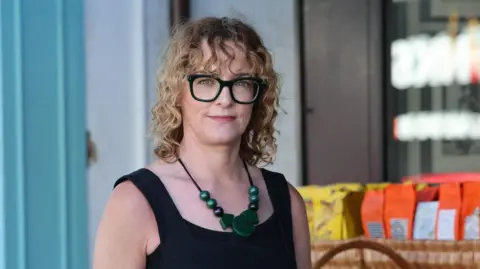 BBC Laura has shoulder length curly blonde hair with a curly fringe. She is wearing black framed glasses and a black vest top. She has a large green necklace on.