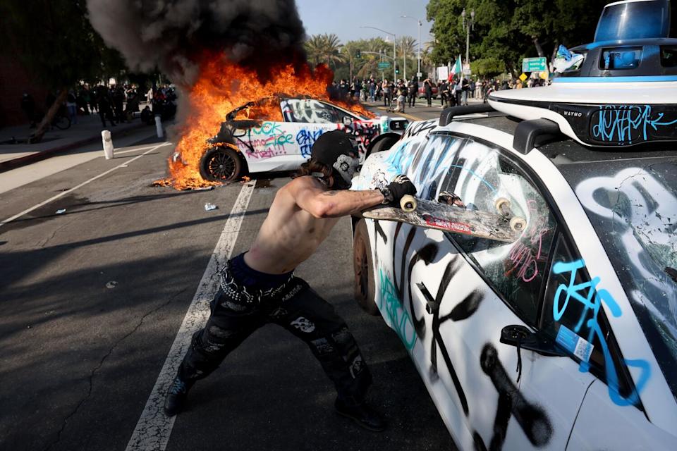 A shirtless man smashes a skateboard into the window of a car as another car burns behind him