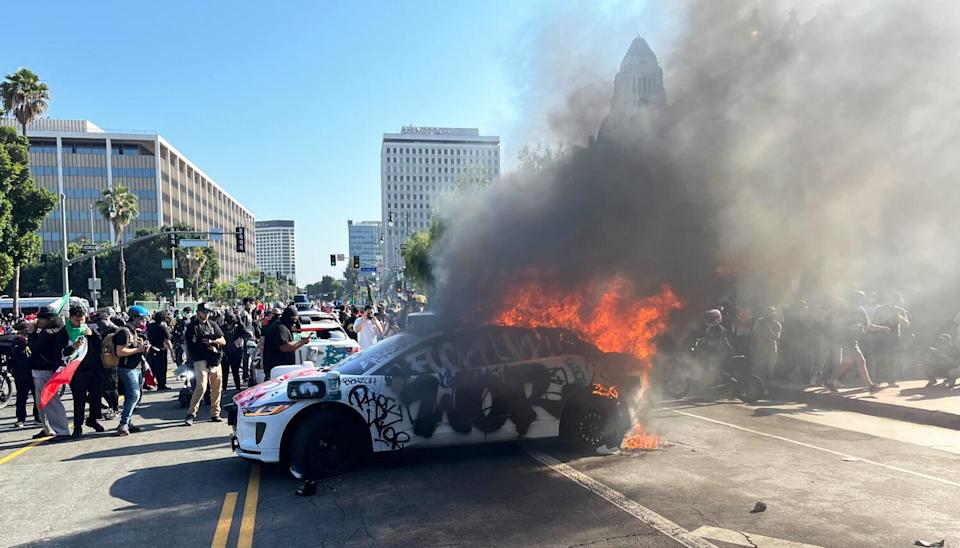 A white vehicle covered in graffiti burns on a street, spewing flames and smoke