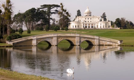 A long shot of Stoke Park house, a white manor with a signature dome. In the foreground is a bridge over a lake. 