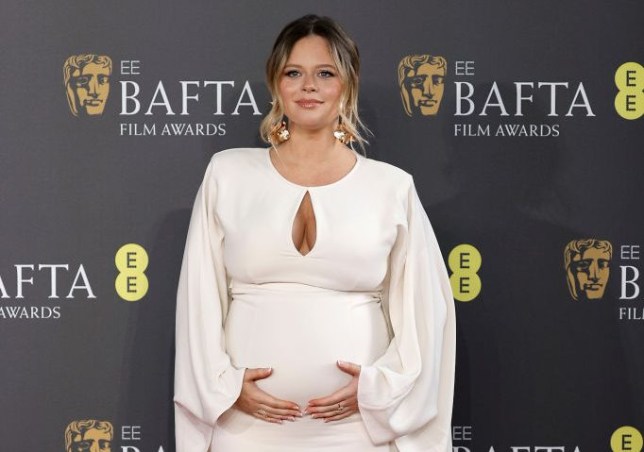 Emily Atack in white gown at 2024 EE BAFTA Film Awards, cradling pregnancy bump