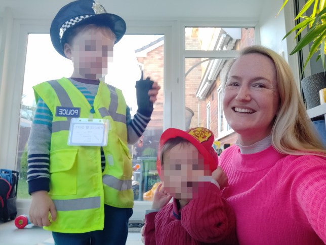 Tarryn in a pink jumper takes a selfie with her kids - dressed as a police man and fire man - both kids faces are blurred