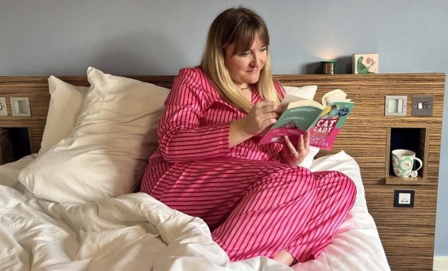 Alison Perry in striped pink pyjamas reading a book in bed