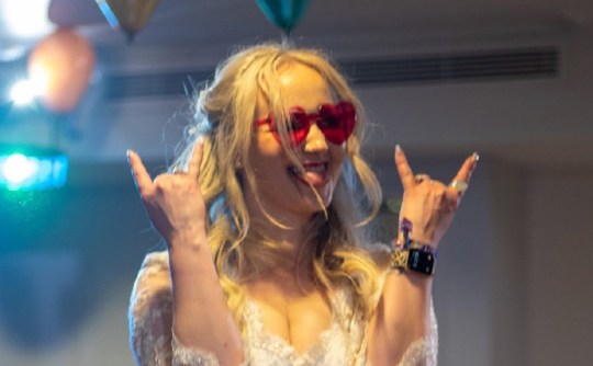 Rebecca wearing heart shaped pink sunglasses, a wedding dress, her tongue out and doing 'rock on' signs with her hands