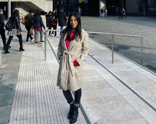 Angela posing in front of a building, on the steps, wearing a trenchcoat with a belt