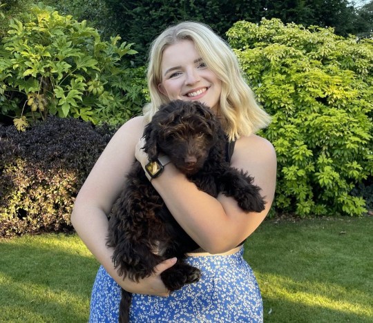 Jess in her garden in a blue dress, holding a dark brown dog and smiling
