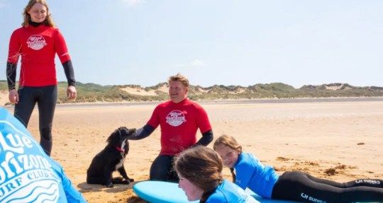 Ariel the six legged mermaid dog with surf teachers and children learning to surf on a beach. 