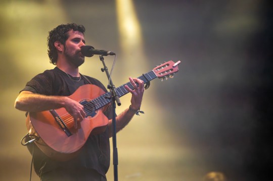 Nick Mulvey with brown hair and a beard strums a light brown guitar as he sings into a microphone while wearing black t-shirt and trousers.