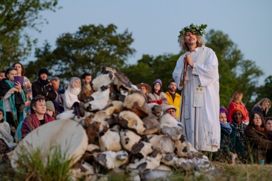 A man with long curly grey hair stands with a stick while wearing a white robe and a leafy crown as the crowd, some seated near the tree line, watch the sun rise.