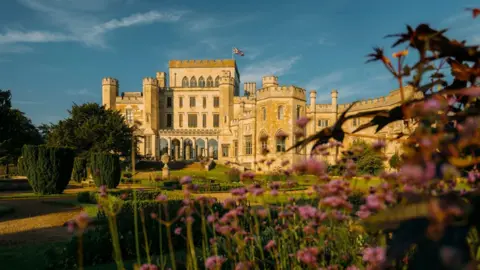 Ashridge House A castellated stone manor house against a blue sky with yew trees, box hedging and pink flowers in front, Ashridge House 