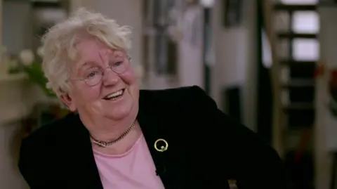 BBC Nell McCafferty in 2016, smiling and looking off camera. She is wearing glasses, a black blazer, pink top and gold broach.