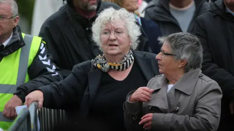 PA Media Nell McCafferty walking and talking with with another woman, her hand on a metal barrier 