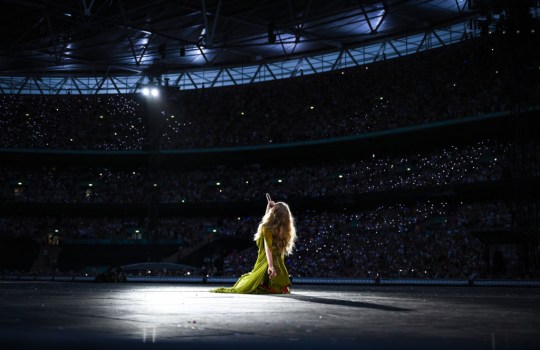 Taylor Swift kneels on ground facing fans as she sings folklore and evermore eras during Eras Tour 