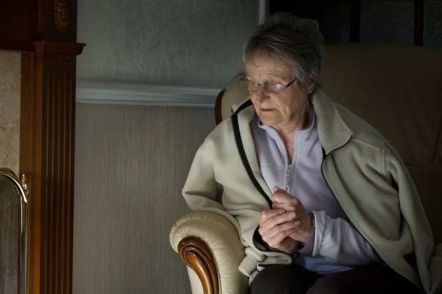 An elderly woman in a dimly lit room wearing a fleece cardigan looking pensively to the side