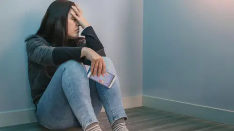 Getty Images Depressed woman sits in the corner of an empty room with her head in her hand and a mobile phone in the other hand (picture posed by a model).