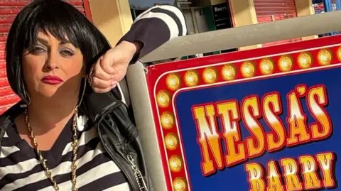 Knock Off Nessa Nessa impersonator Kaz Barrett in full get up next to a sign for Nessa's Slots in Barry Island